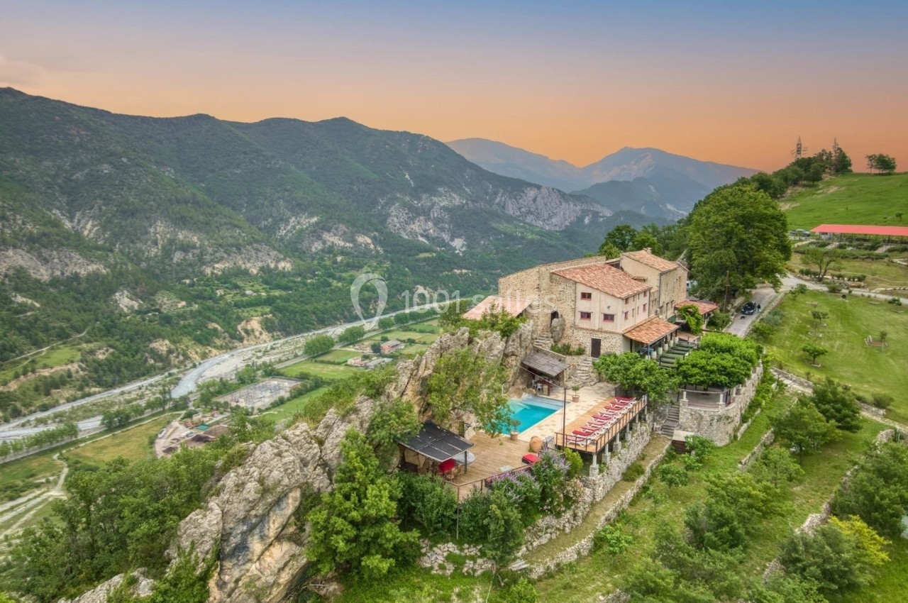 Vue aérienne d'un bâtiment en pierre avec piscine, perché sur une colline verdoyante, entouré de montagnes au coucher du…