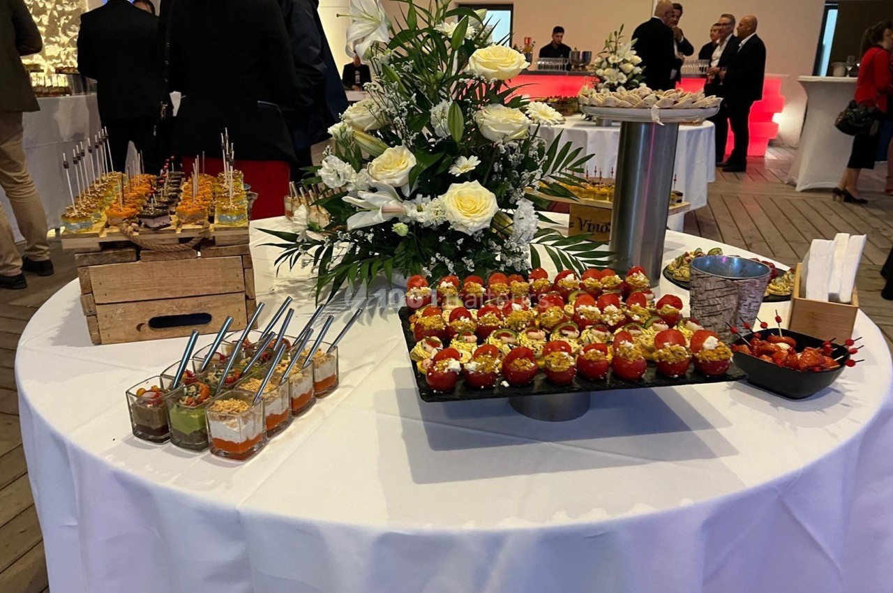 Table ronde avec buffet de verrines, fleurs blanches en centre de table, et invités en arrière-plan.