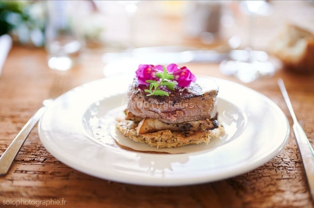 Assiette avec un filet de viande sur un lit de légumes et une garniture florale, servie sur une table en bois.