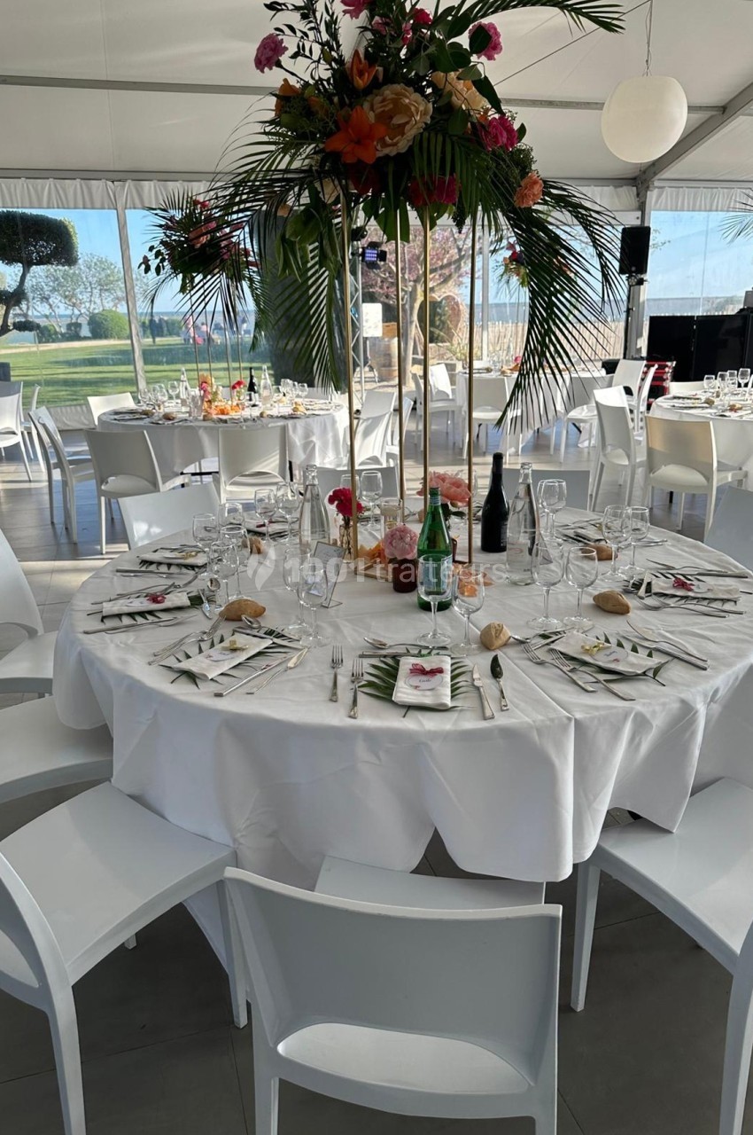 Table ronde élégamment dressée avec nappes blanches, vaisselle raffinée et centre de table floral dans une salle lumineuse.