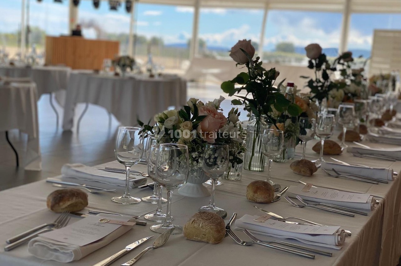 Table élégamment dressée avec nappes blanches, verres, couverts, menus, fleurs et petits pains dans une salle lumineuse.