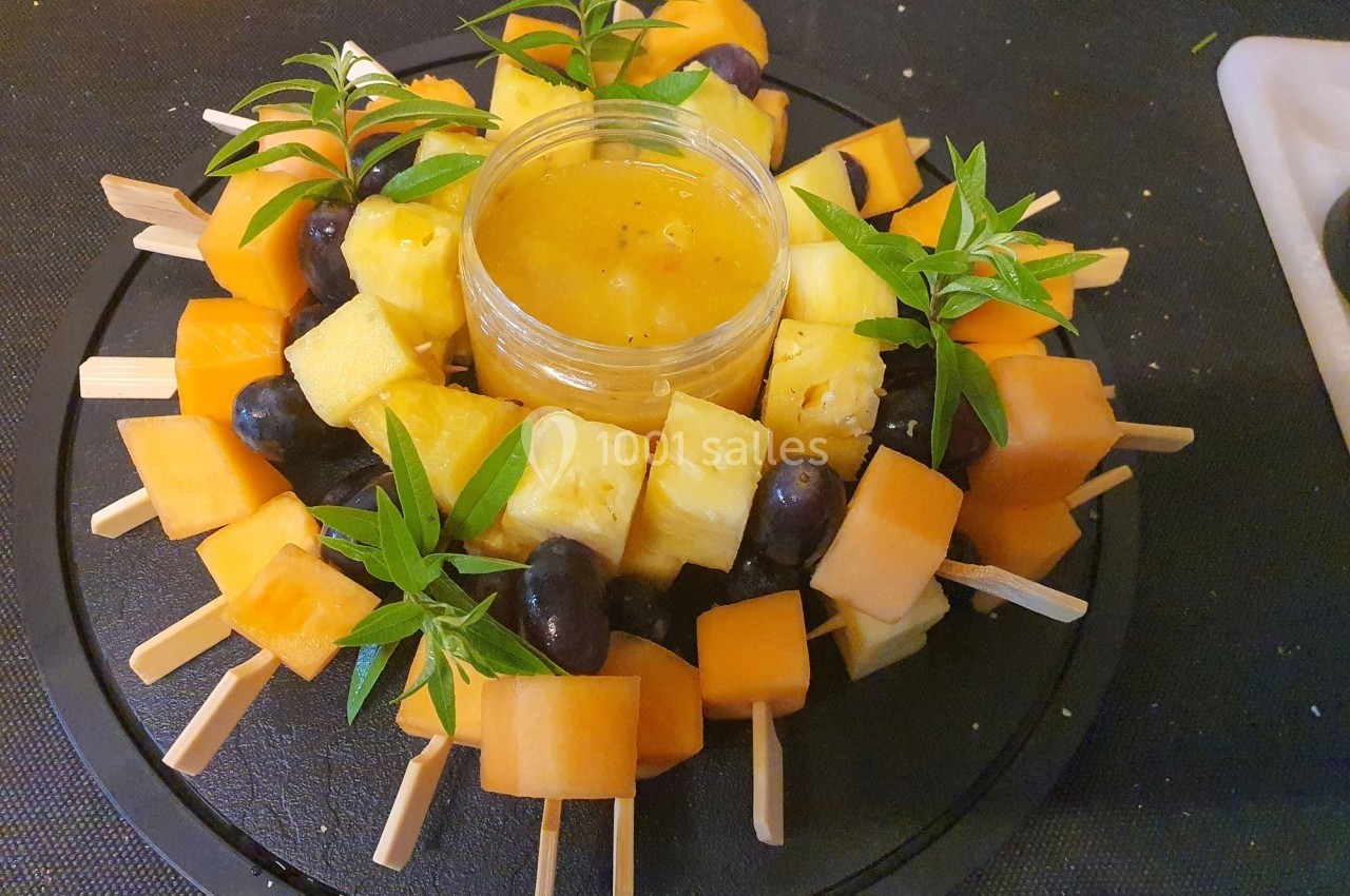 Brochettes de fruits variés avec ananas, melon et raisins autour d'un pot de sauce au centre, décorées de feuilles vertes.