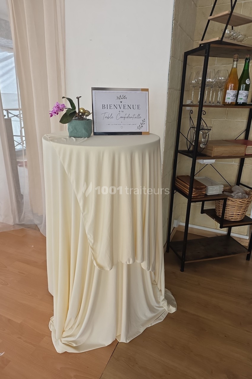 Table haute recouverte d'une nappe beige, décorée d'une orchidée et d'un cadre ’Bienvenue’, près d'une étagère.