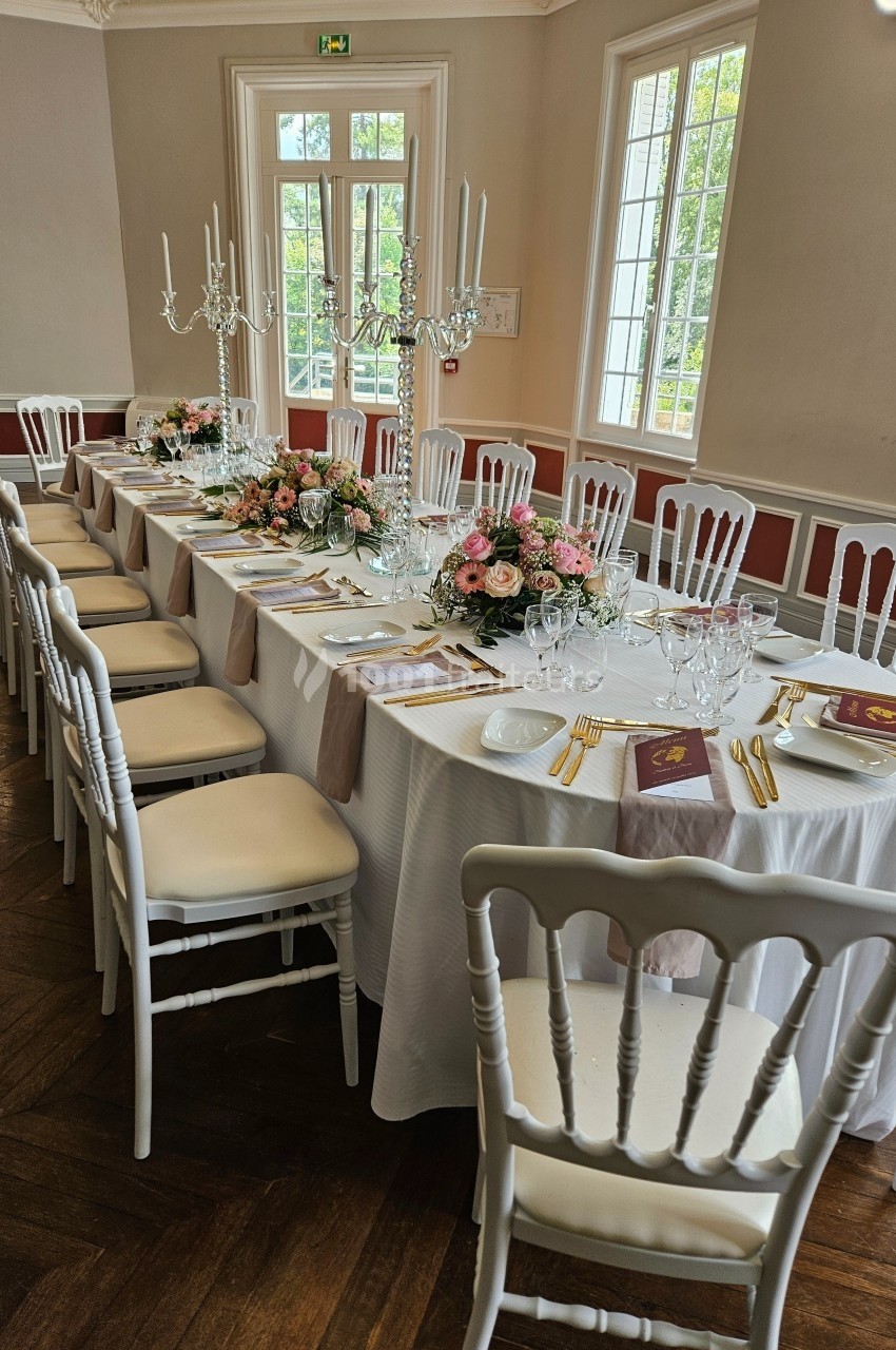 Table élégamment dressée avec nappes blanches, chandeliers, fleurs roses et vaisselle raffinée dans une salle lumineuse.