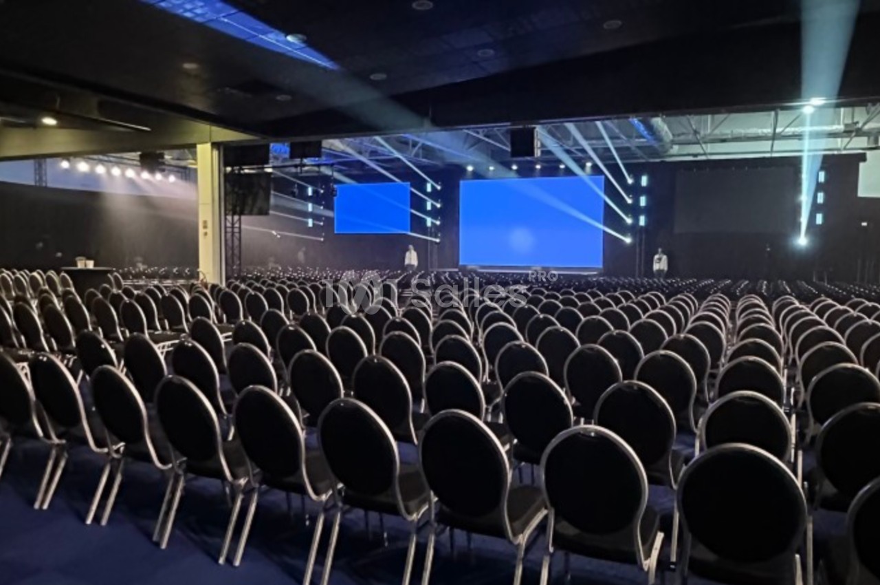 Salle de conférence vide avec rangées de chaises noires alignées face à une grande scène équipée d'écrans.