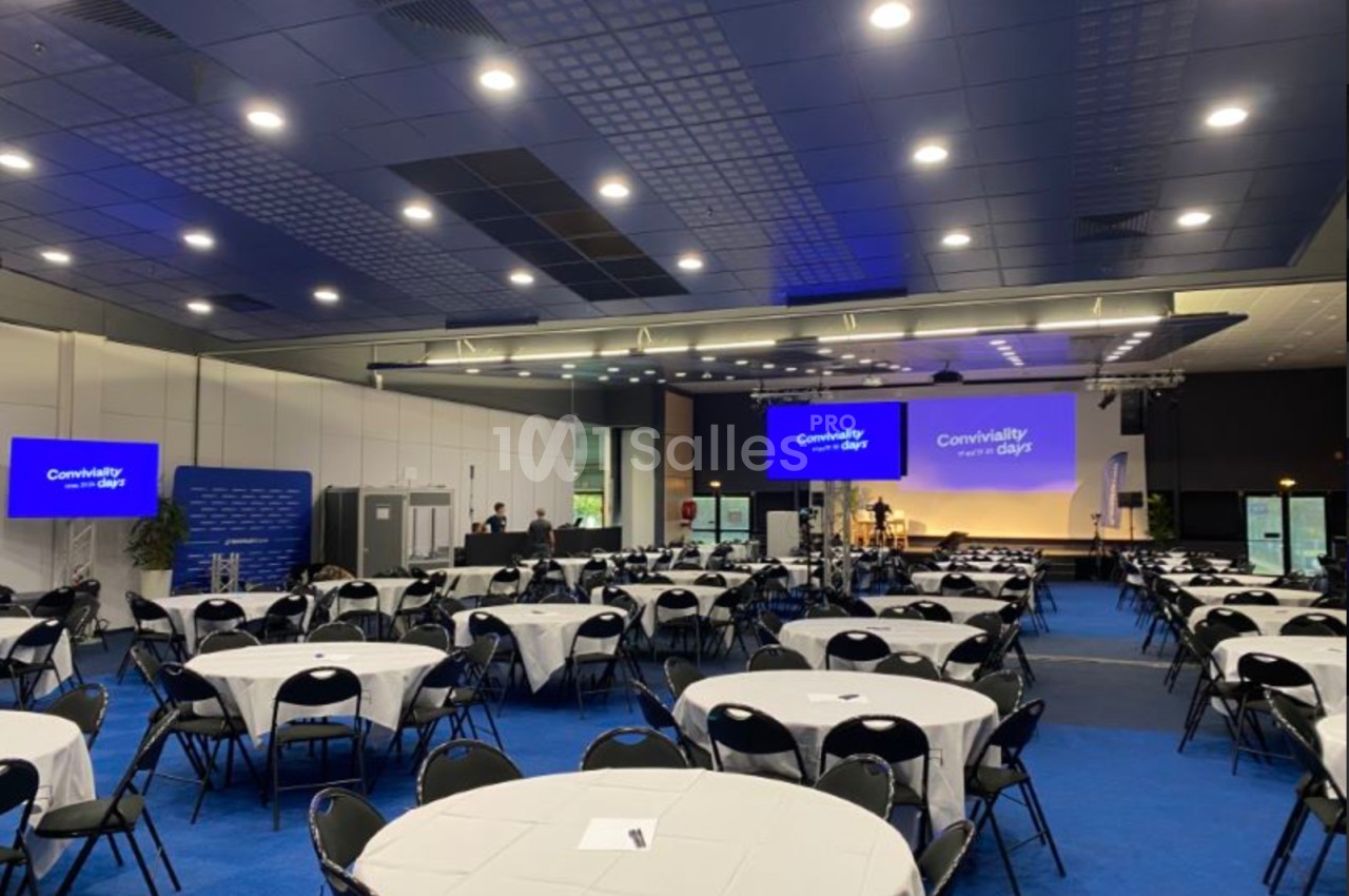 Salle de conférence avec tables rondes dressées, écrans affichant ’Conviality at heart days’ et éclairage moderne.