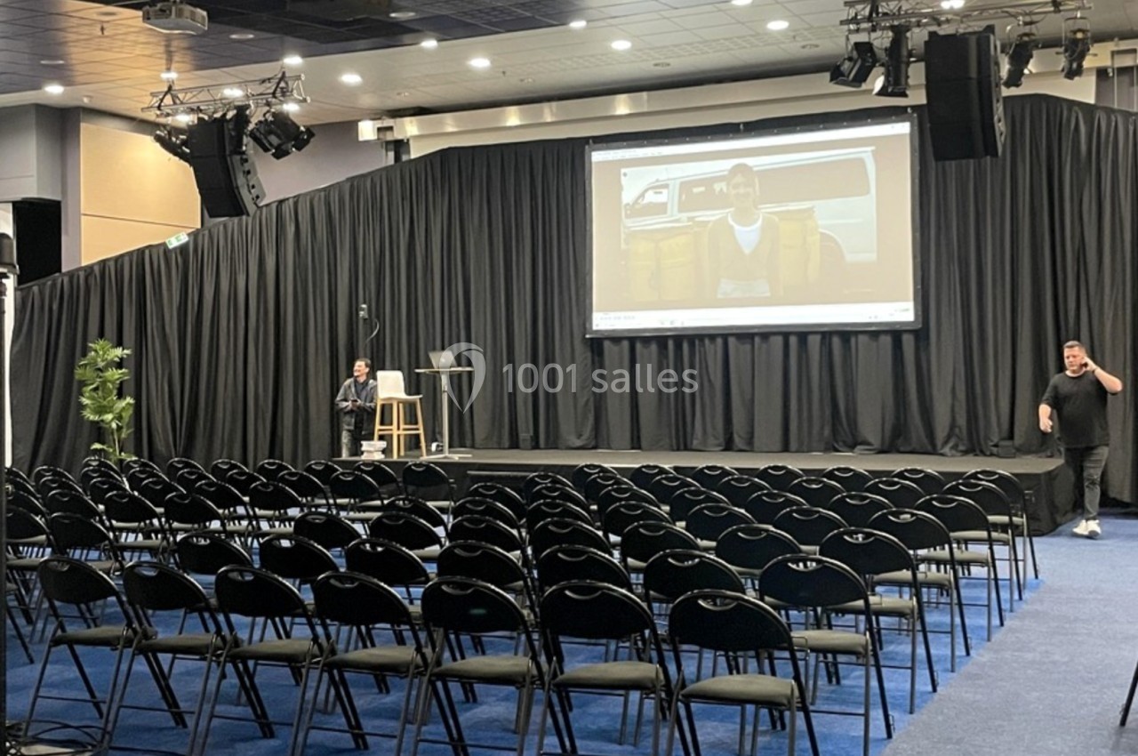 Salle de conférence avec des rangées de chaises vides, une scène équipée d'un écran géant et deux personnes présentes.