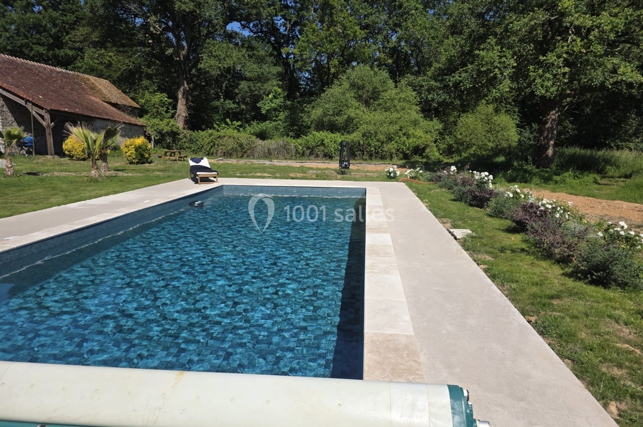 Piscine extérieure entourée de verdure, avec margelles en pierre et transat sur la terrasse.
