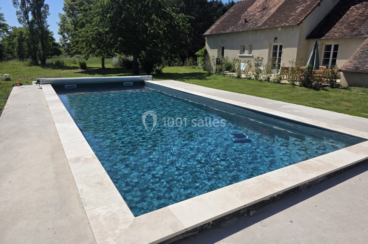 Piscine rectangulaire avec eau claire, entourée de margelles en pierre, située près d'une maison à la campagne.