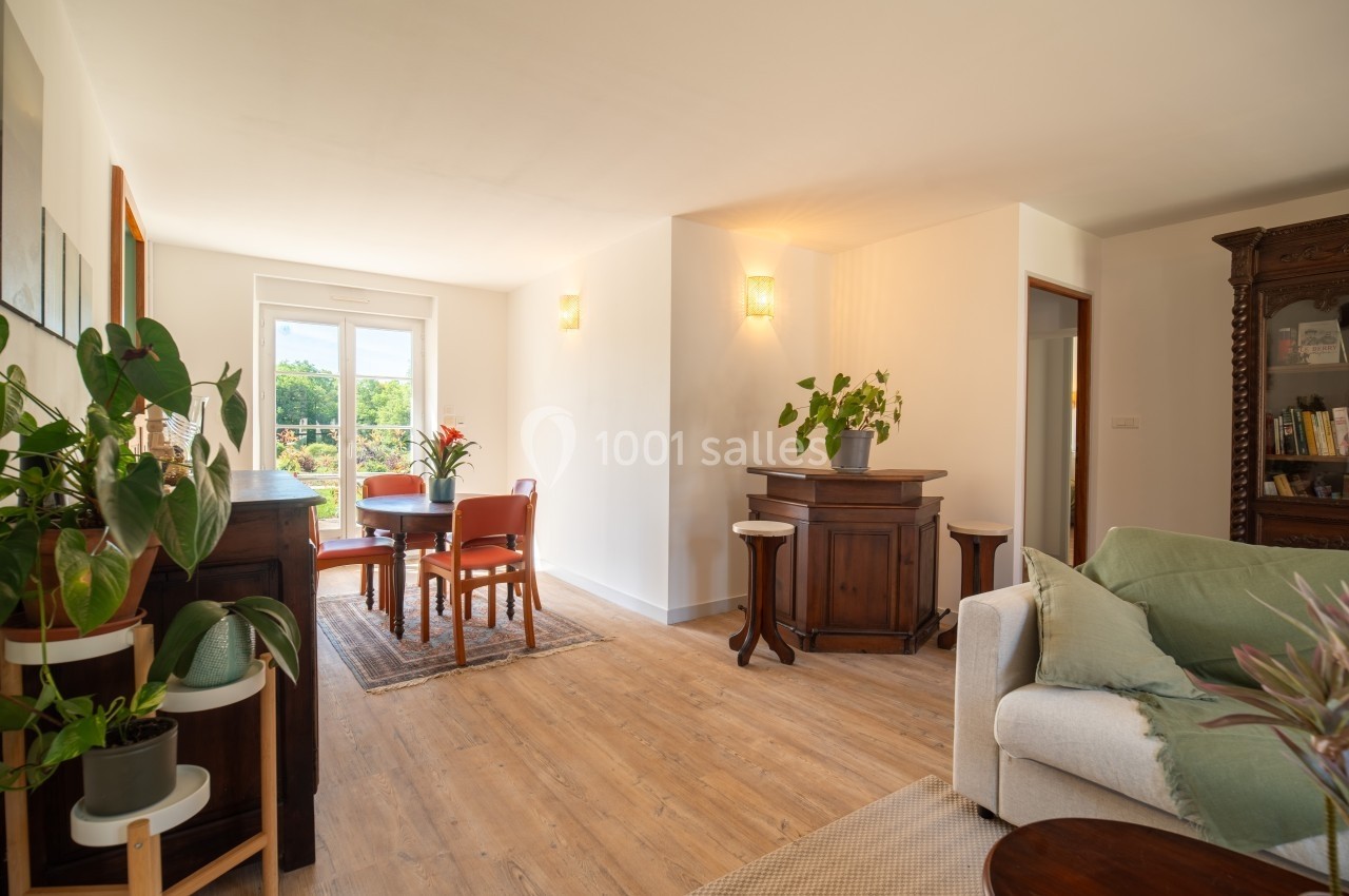 Salon lumineux avec parquet, canapé, meubles en bois, plantes et table à manger près d'une fenêtre donnant sur l'extérieur.