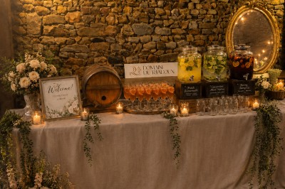 Salle en pierre avec poutres apparentes, tables décorées pour un dîner, invités debout et assis en arrière-plan.