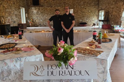 Salle en pierre avec poutres apparentes, tables décorées pour un dîner, invités debout et assis en arrière-plan.