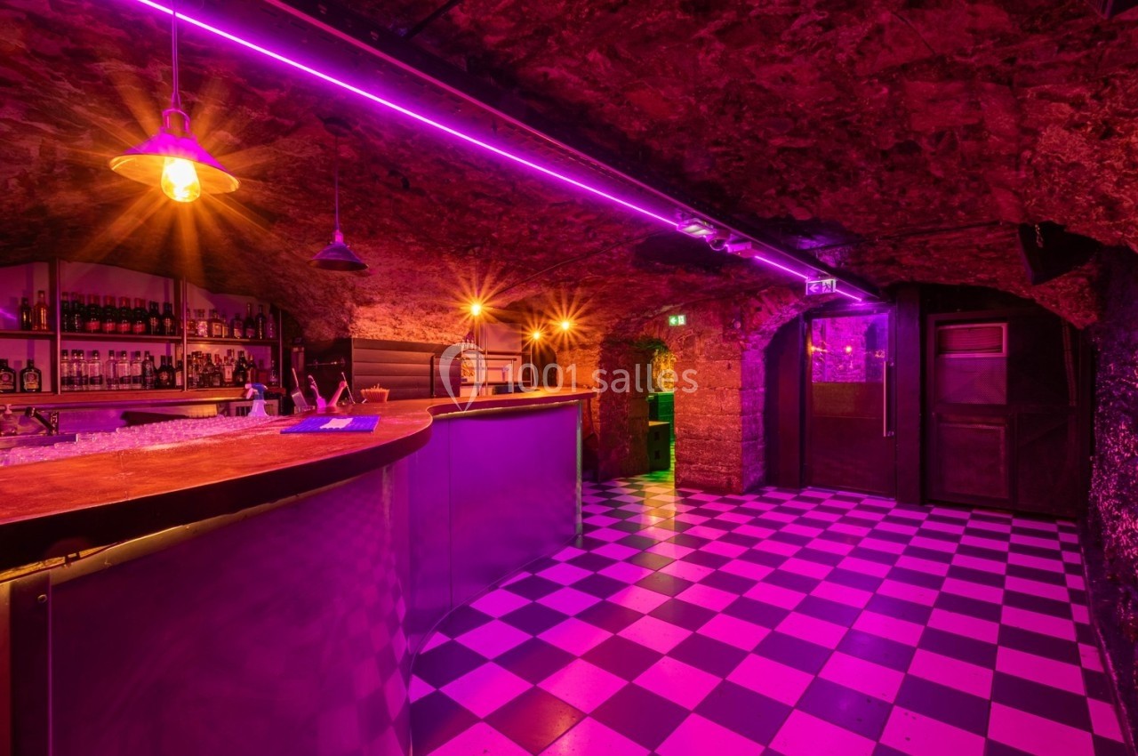 Bar souterrain avec éclairage violet, comptoir en bois, étagères de bouteilles et sol à carreaux noir et blanc.