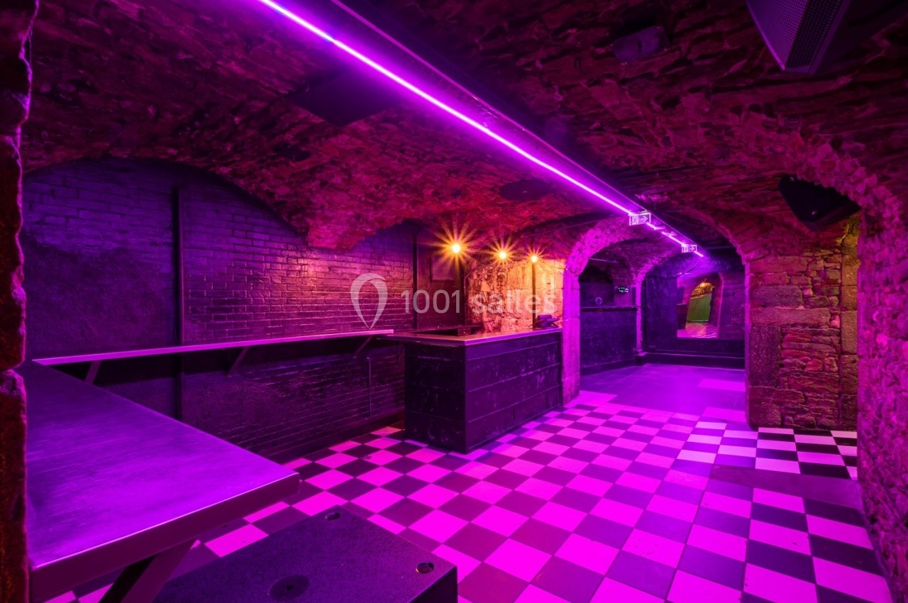 Salle voûtée en pierre avec éclairage violet, sol en damier noir et blanc, et comptoir central.