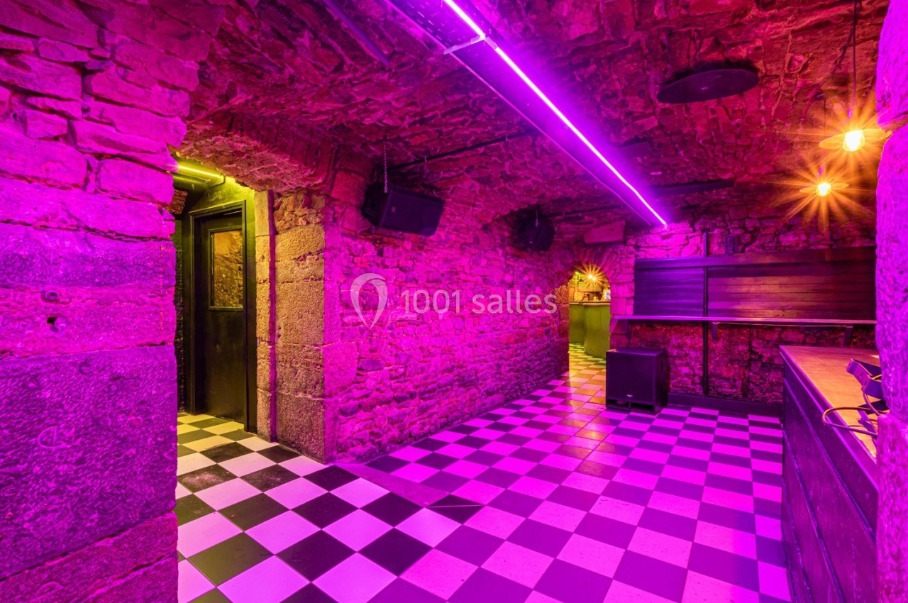 Salle en sous-sol avec murs en pierre, éclairage néon violet et sol à carreaux noirs et blancs.