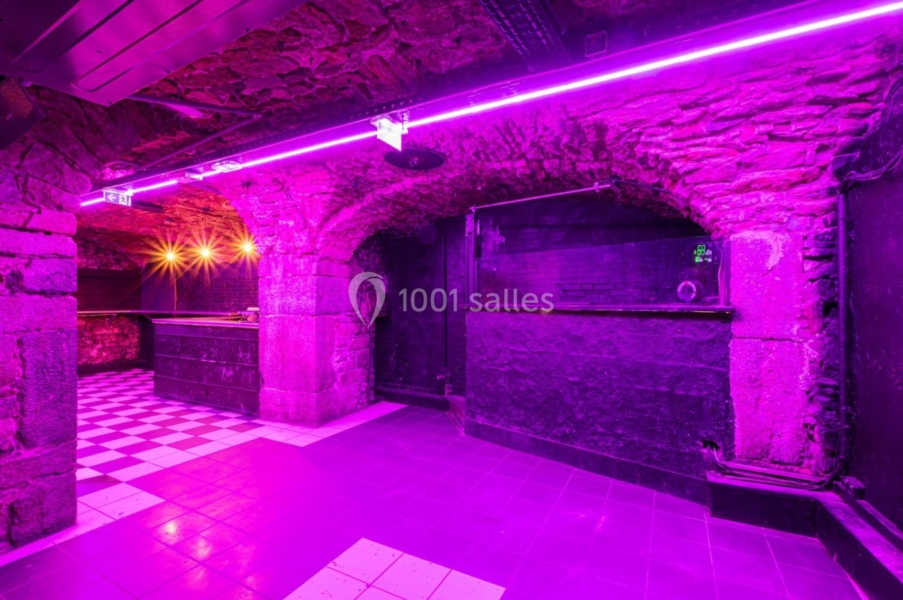 Salle voûtée en pierre avec éclairage violet, sol carrelé et comptoir sombre dans un espace intérieur.