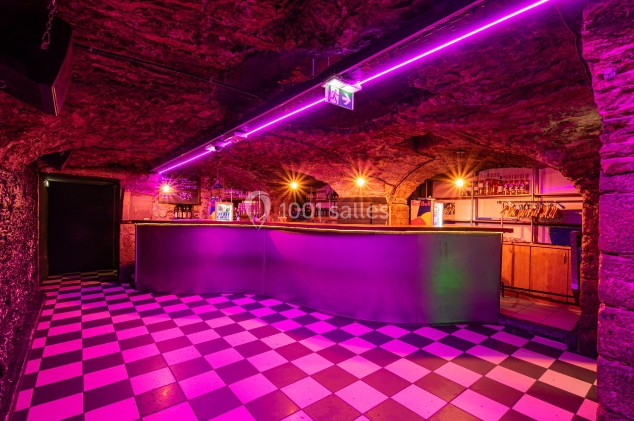 Bar souterrain avec comptoir éclairé par des néons violets, sol en damier noir et blanc et murs en pierre.