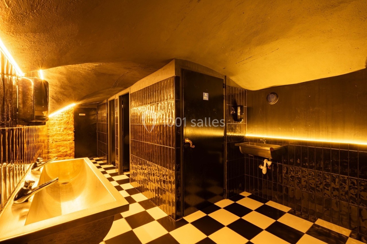 Salle de bain au design moderne avec murs noirs, éclairage chaleureux, lavabo doré et sol en damier noir et blanc.