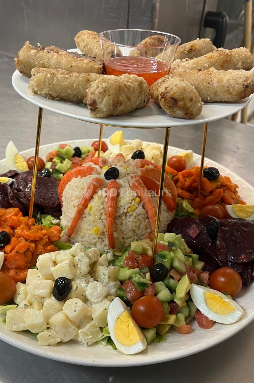 Assiette garnie de crudités, riz, crevettes et œufs, surmontée d'un plateau de nems avec sauce rouge.