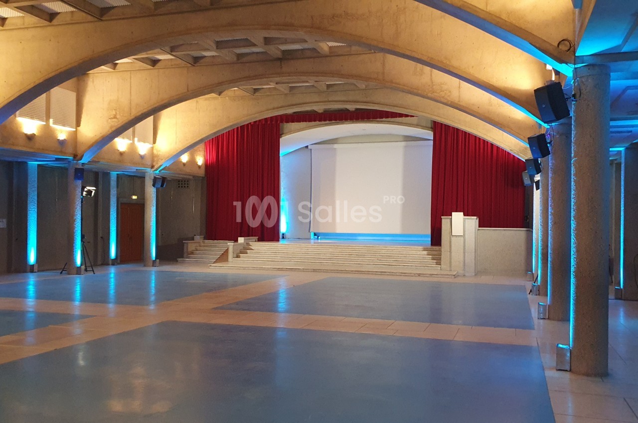 Grande salle vide aux voûtes en béton apparentes, avec estrade surélevée, rideaux rouges, écran de projection et colonnes éclairées en bleu.
