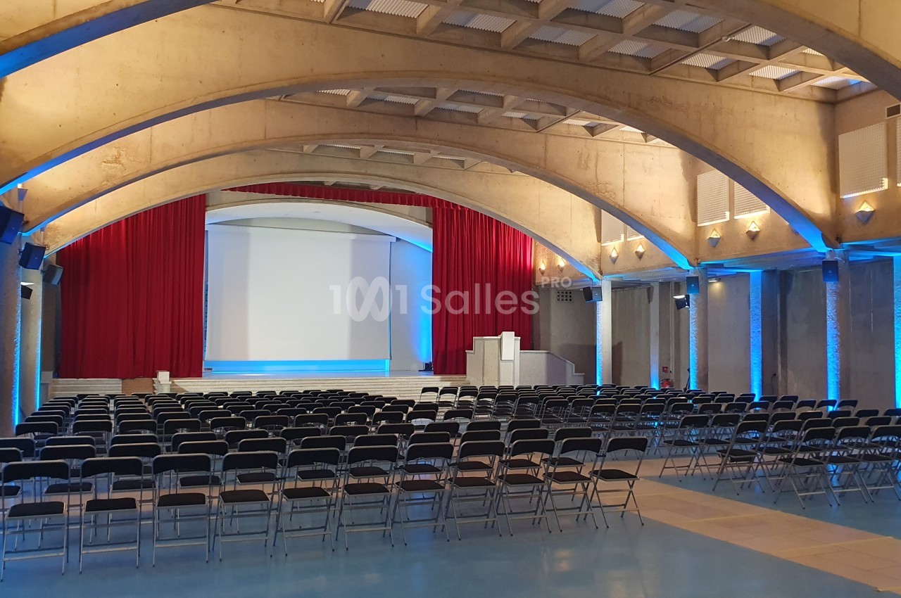 Grande salle à voûtes en béton avec rangées de chaises pliantes face à une scène équipée d'un écran de projection, de rideaux rouges et d'un pupitre.