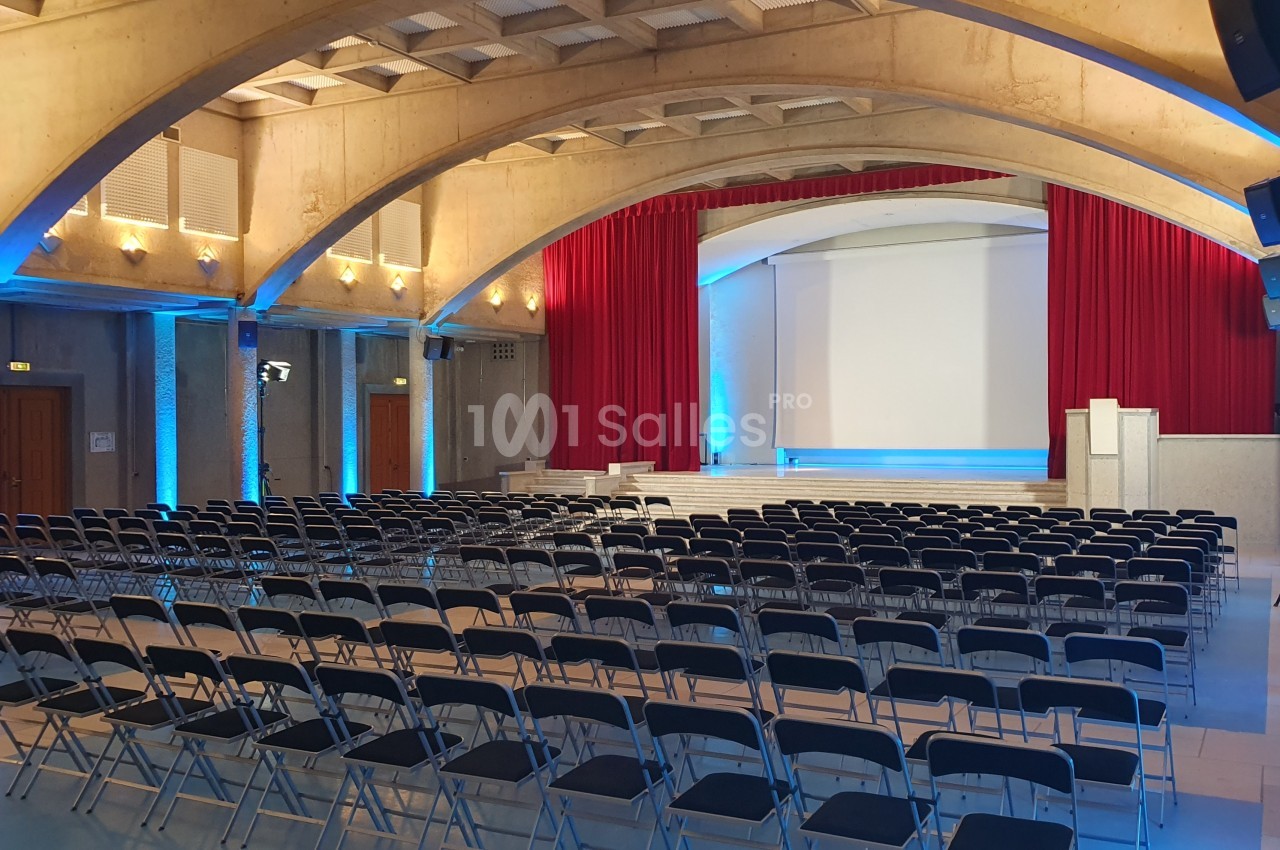 Grande salle de conférence en rangées de chaises pliantes face à une scène avec écran de projection, rideaux rouges et voûtes en béton apparent.
