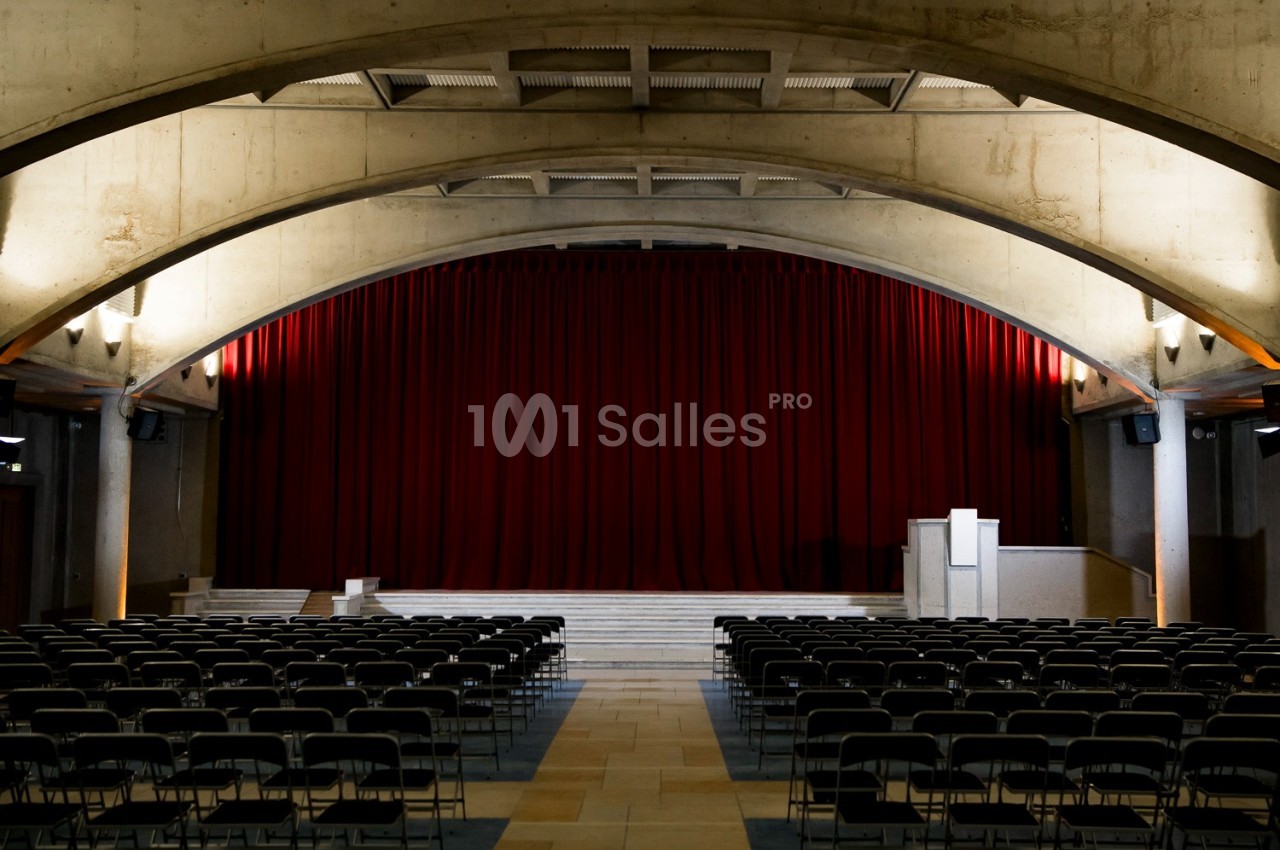Salle de spectacle en intérieur, avec rangées de chaises pliantes face à une scène surélevée et un grand rideau rouge sous des voûtes en béton.