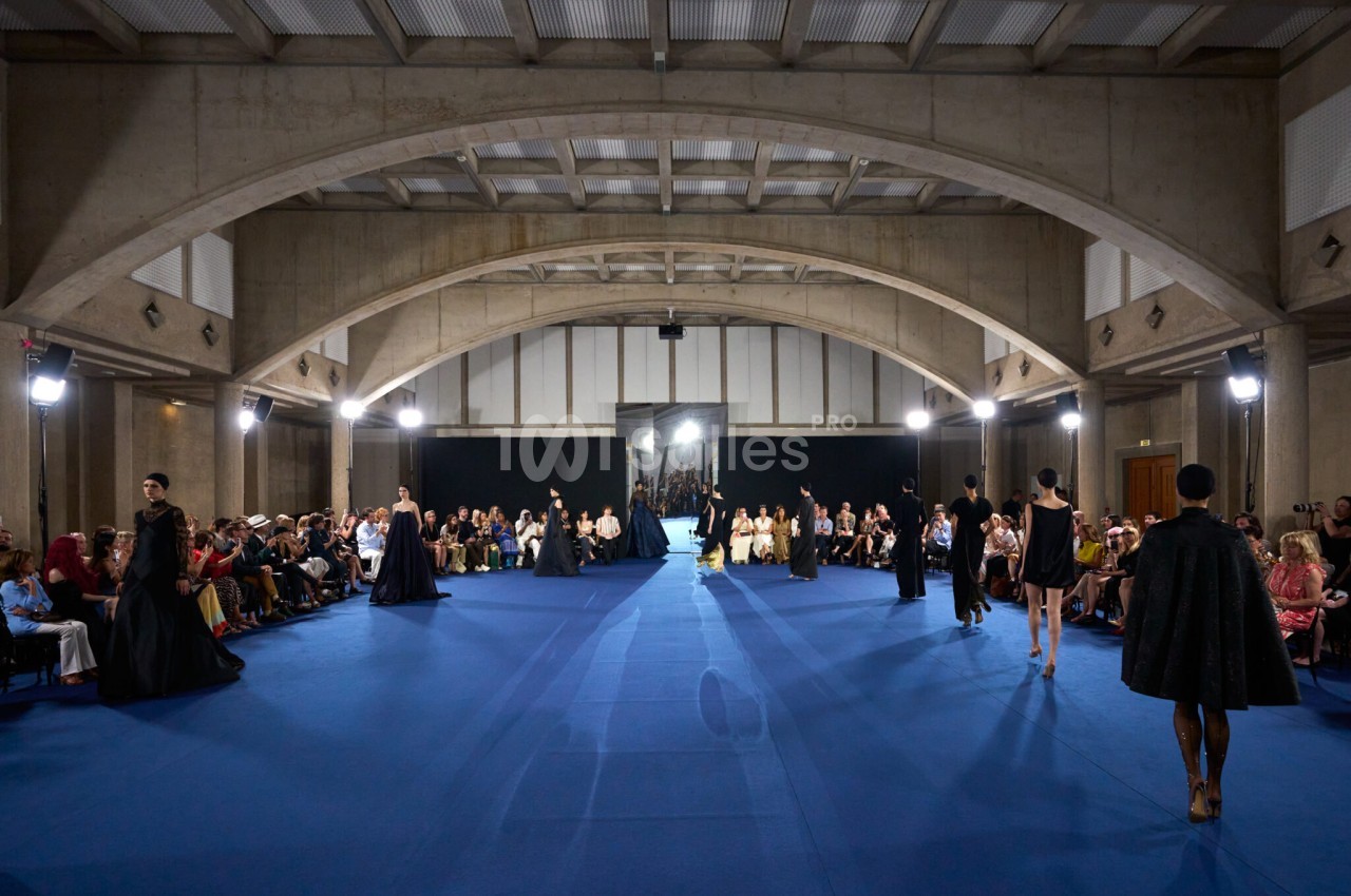Défilé de mode dans une grande salle à voûtes en béton, sur un tapis bleu, avec mannequins en robes et tenues noires et public assis de part et d'autre.