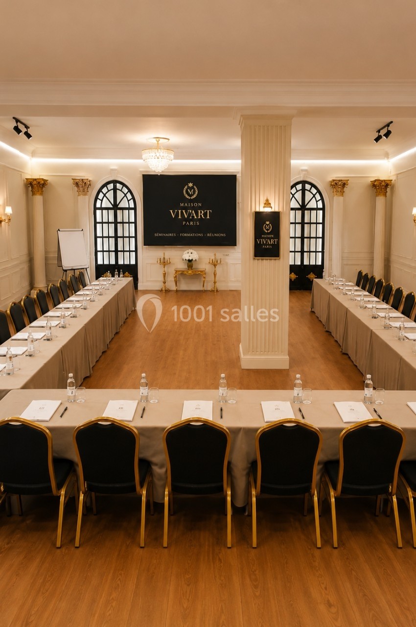 Salle de conférence élégante avec tables en U, chaises noires, écran, paperboards et décoration raffinée.