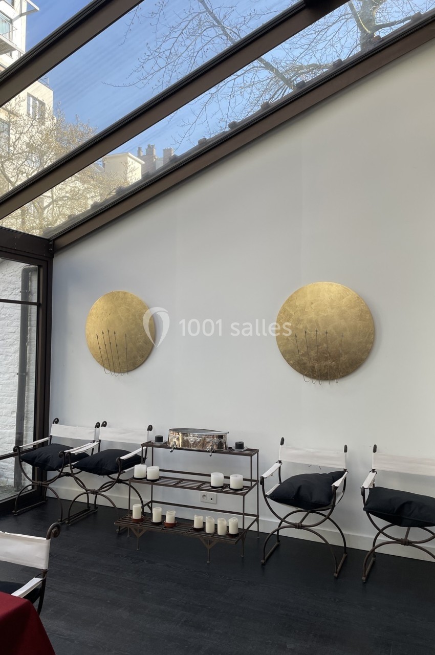 Salle lumineuse avec verrière, murs blancs décorés de disques dorés, chaises design et étagère avec bougies.