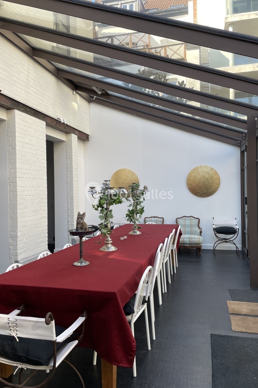 Salle à manger lumineuse avec une grande table recouverte d'une nappe rouge, entourée de chaises blanches, sous une verrière.