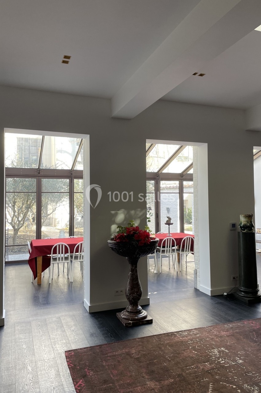 Salle lumineuse avec grandes baies vitrées, tables dressées avec nappes rouges et vue sur un jardin extérieur.