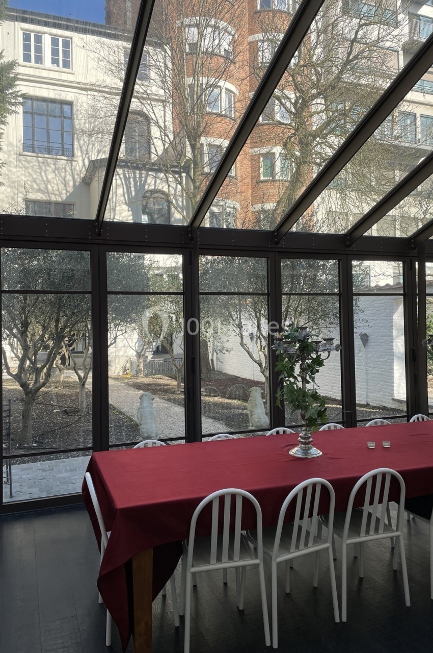 Salle vitrée donnant sur un jardin avec des oliviers, une grande table rouge et des chaises blanches au premier plan.