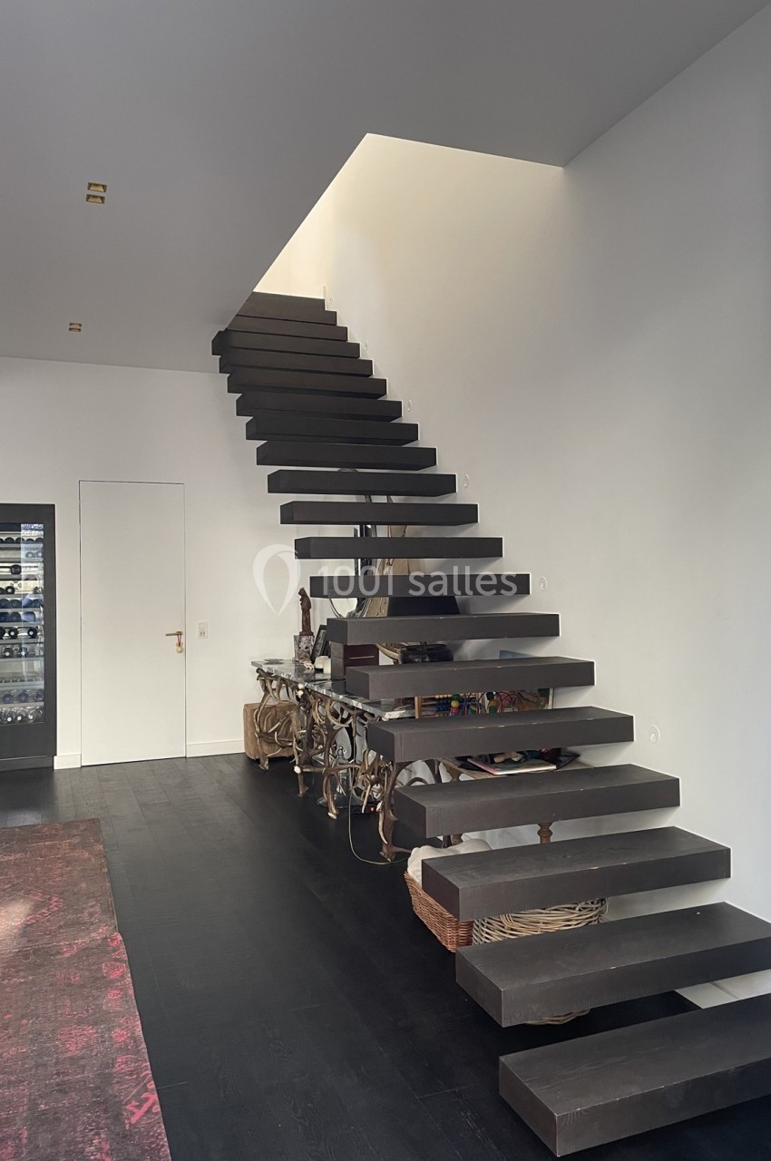 Escalier suspendu en bois sombre dans un intérieur moderne avec murs blancs et sol en parquet foncé.