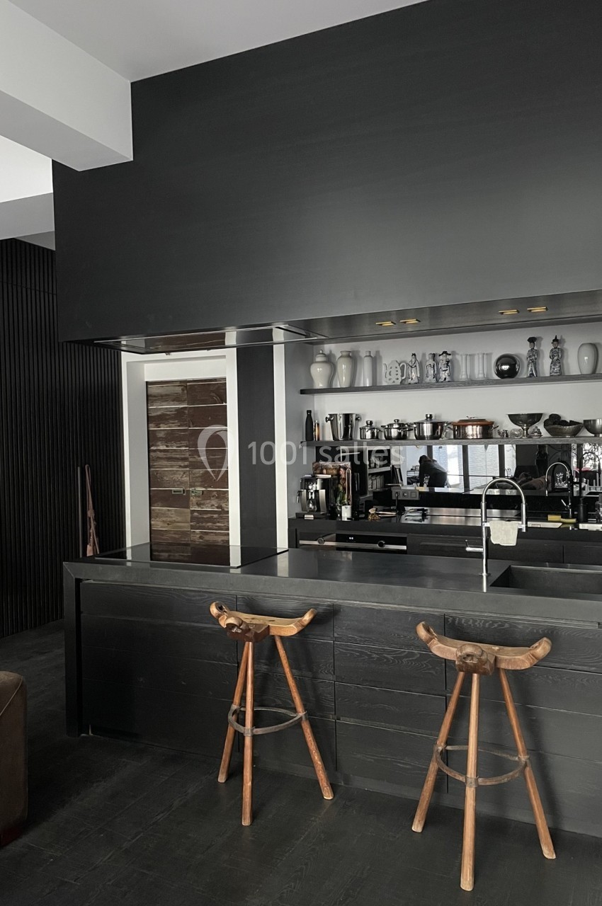 Cuisine moderne avec un comptoir noir, deux tabourets en bois, étagères avec vaisselle et un évier intégré.
