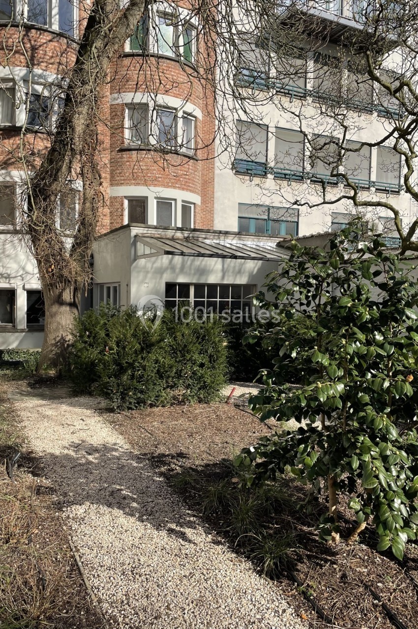 Chemin en gravier bordé de végétation menant à un bâtiment en briques et en crépi avec de grandes fenêtres.