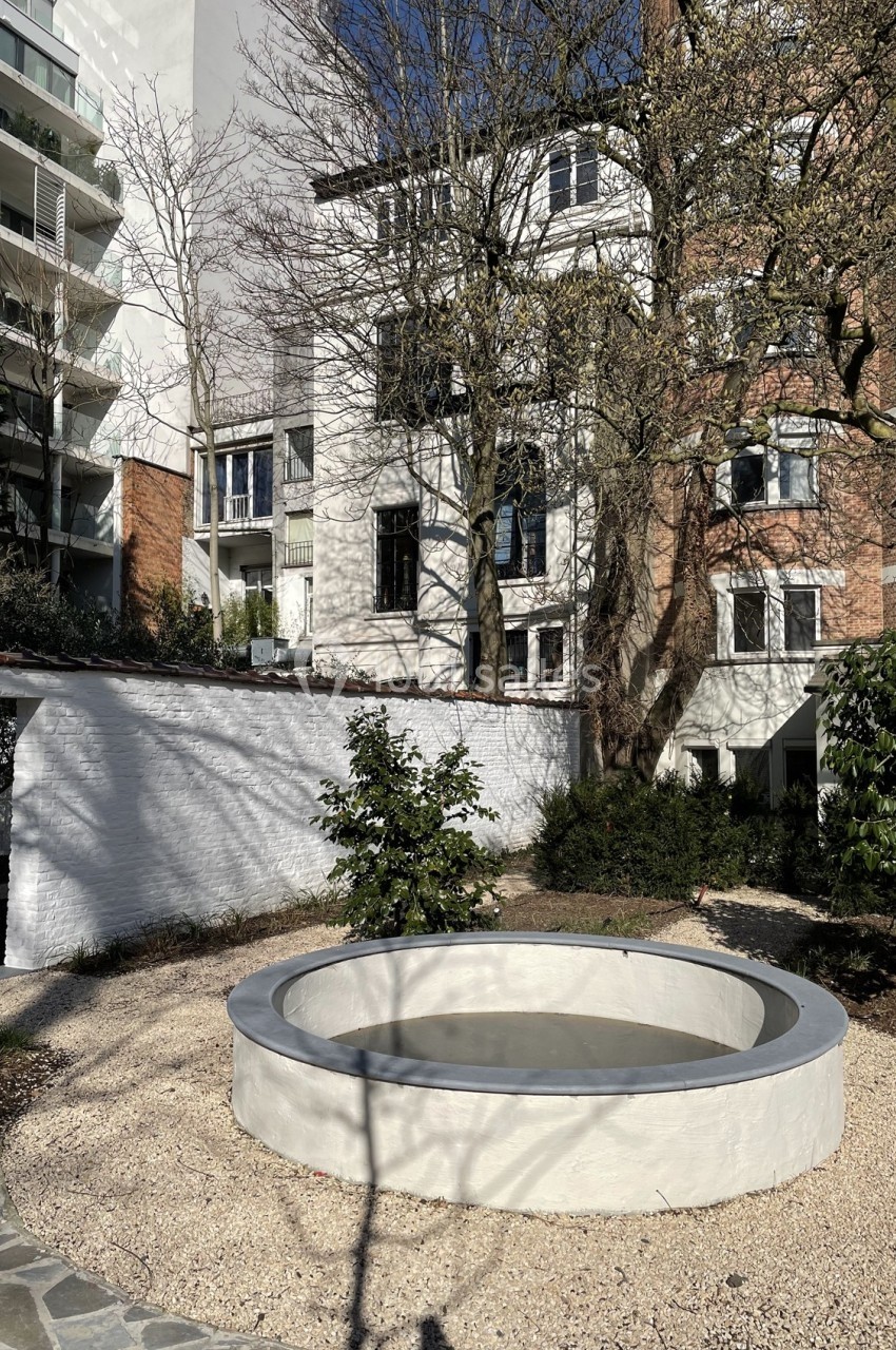 Petit bassin circulaire en béton entouré de gravier, situé dans une cour avec des arbres et des bâtiments en arrière-plan.