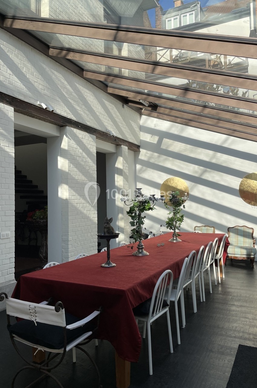Salle à manger lumineuse avec une grande table rouge, des chaises blanches et un toit en verre laissant entrer la lumière…
