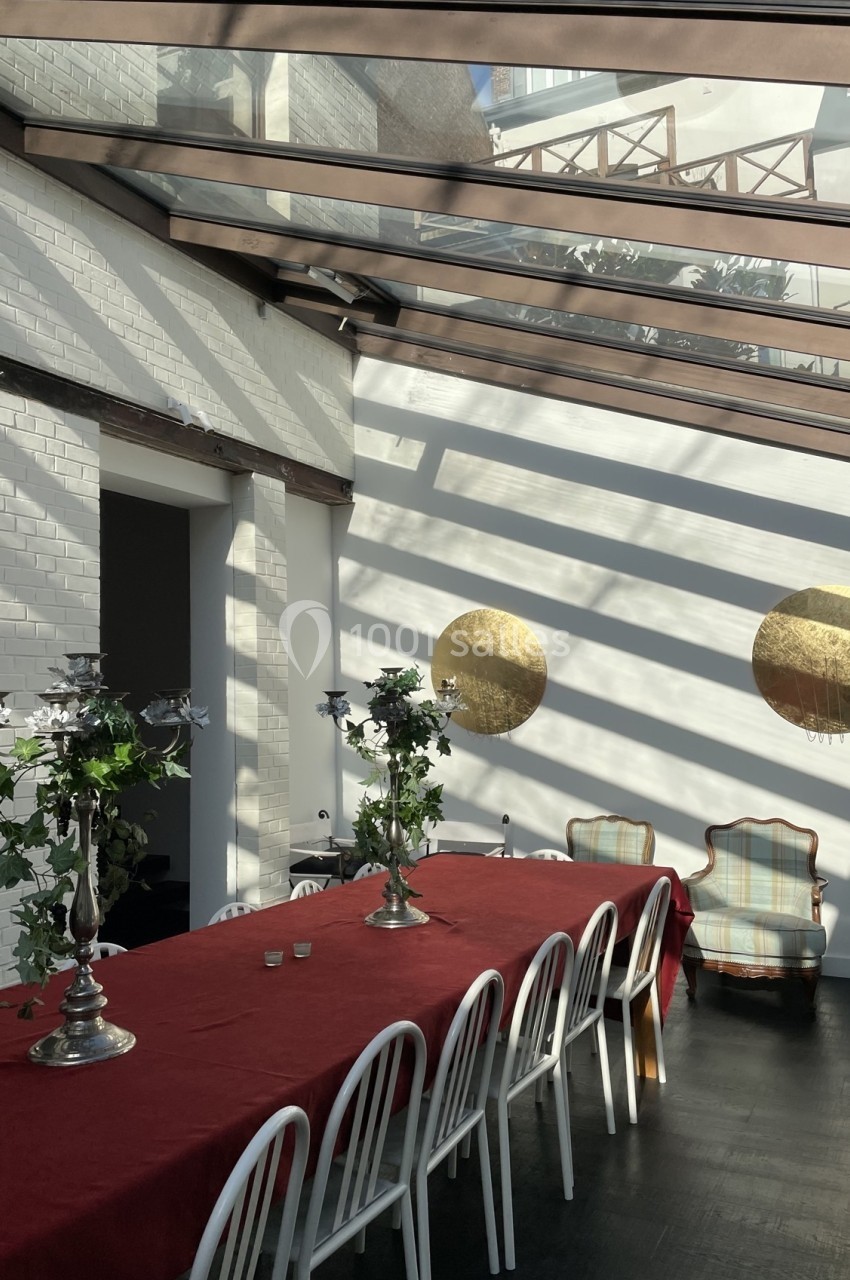 Table longue avec nappe rouge, chaises blanches, chandeliers décorés de feuillage, sous une verrière lumineuse.