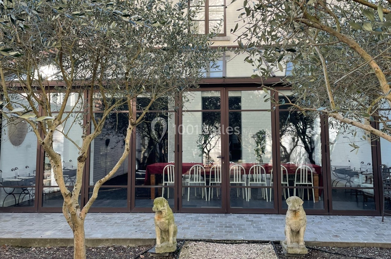 Façade vitrée d'un bâtiment donnant sur une terrasse avec des arbres et deux statues de chiens en pierre.