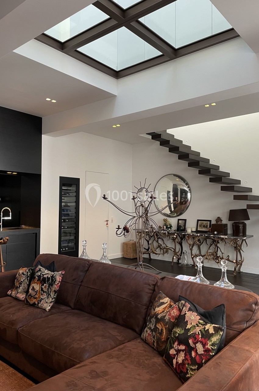 Salon moderne avec canapé en velours marron, coussins à motifs, escalier flottant et grande verrière au plafond.