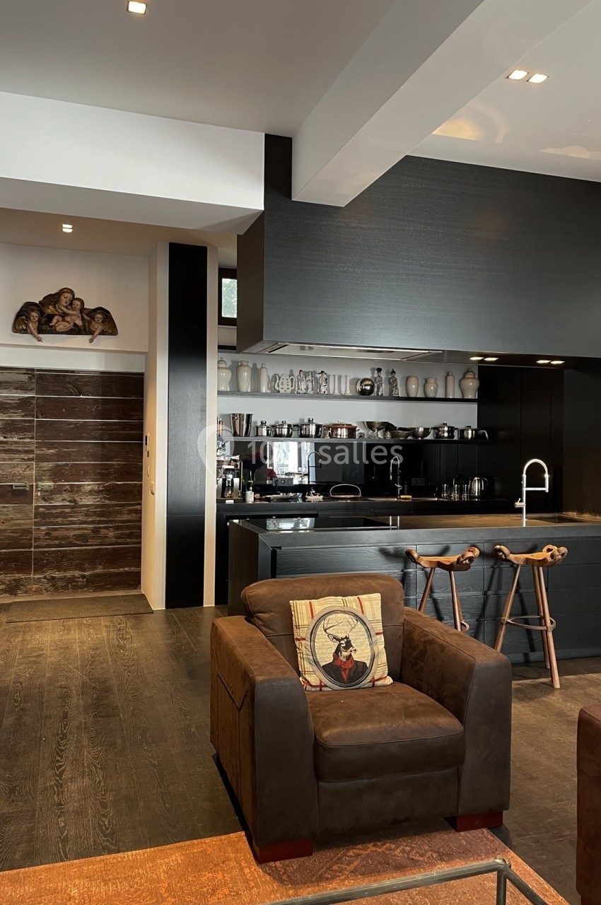 Cuisine moderne ouverte avec comptoir noir, tabourets en bois, étagères exposant vaisselle et salon adjacent avec fauteuil.