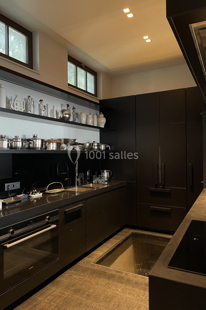 Cuisine moderne avec meubles noirs, étagères ouvertes contenant des ustensiles et lumière naturelle provenant des fenêtres…
