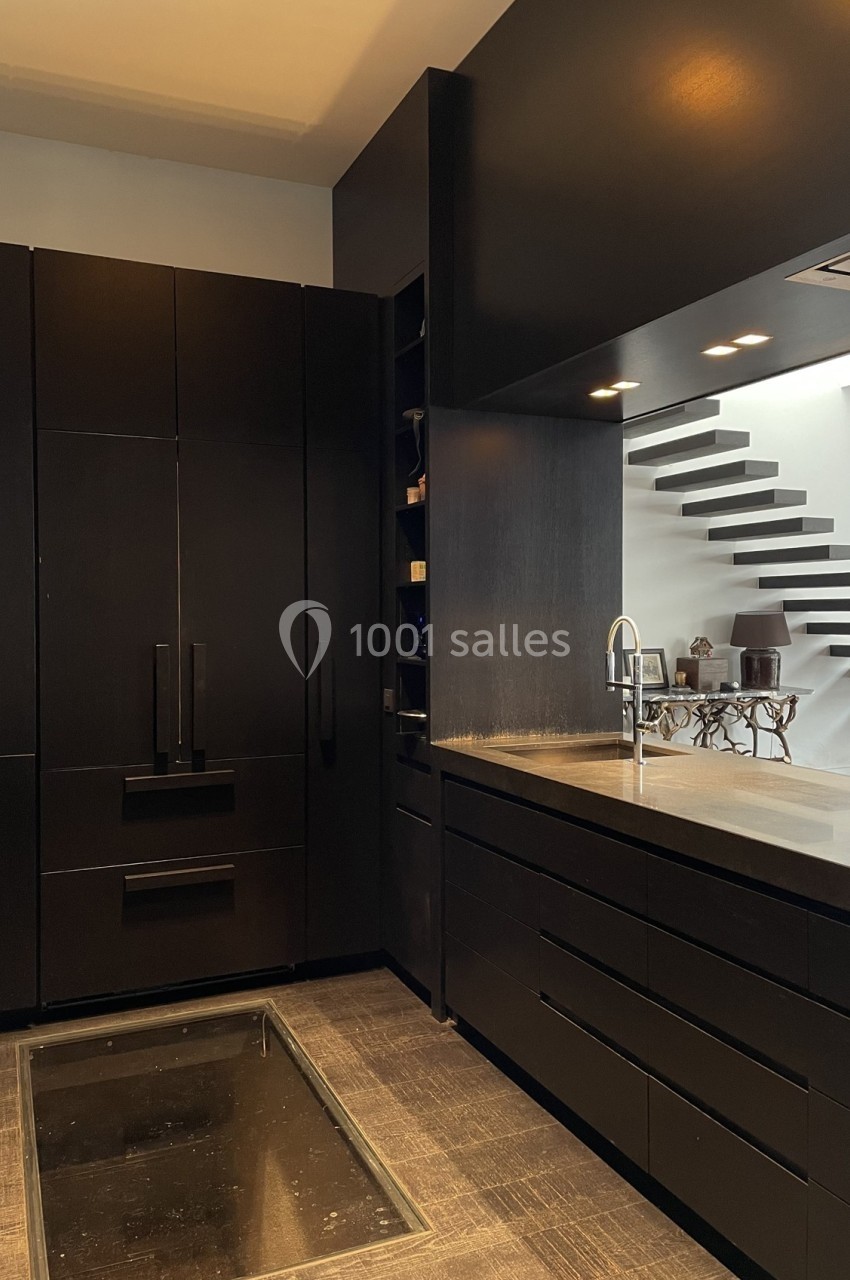 Cuisine moderne avec meubles noirs, plan de travail sombre, évier en inox et escalier minimaliste en arrière-plan.