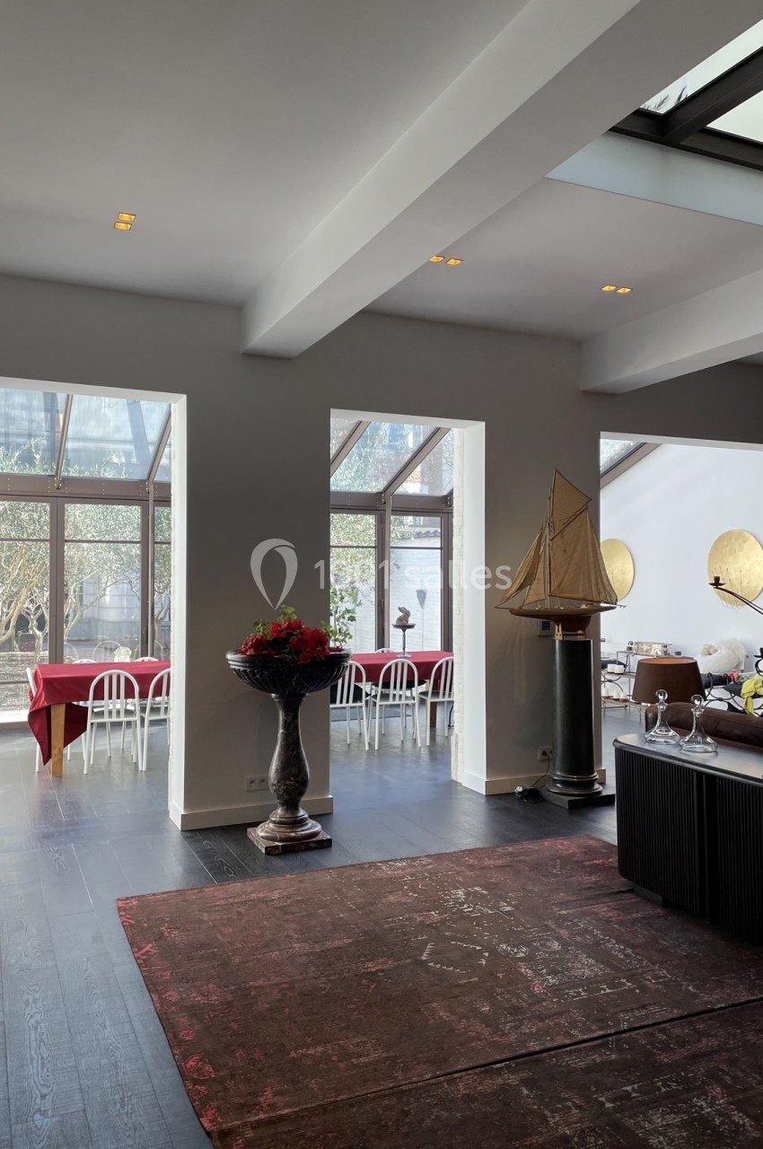 Salle lumineuse avec grandes baies vitrées, tables dressées en arrière-plan et décoration élégante.