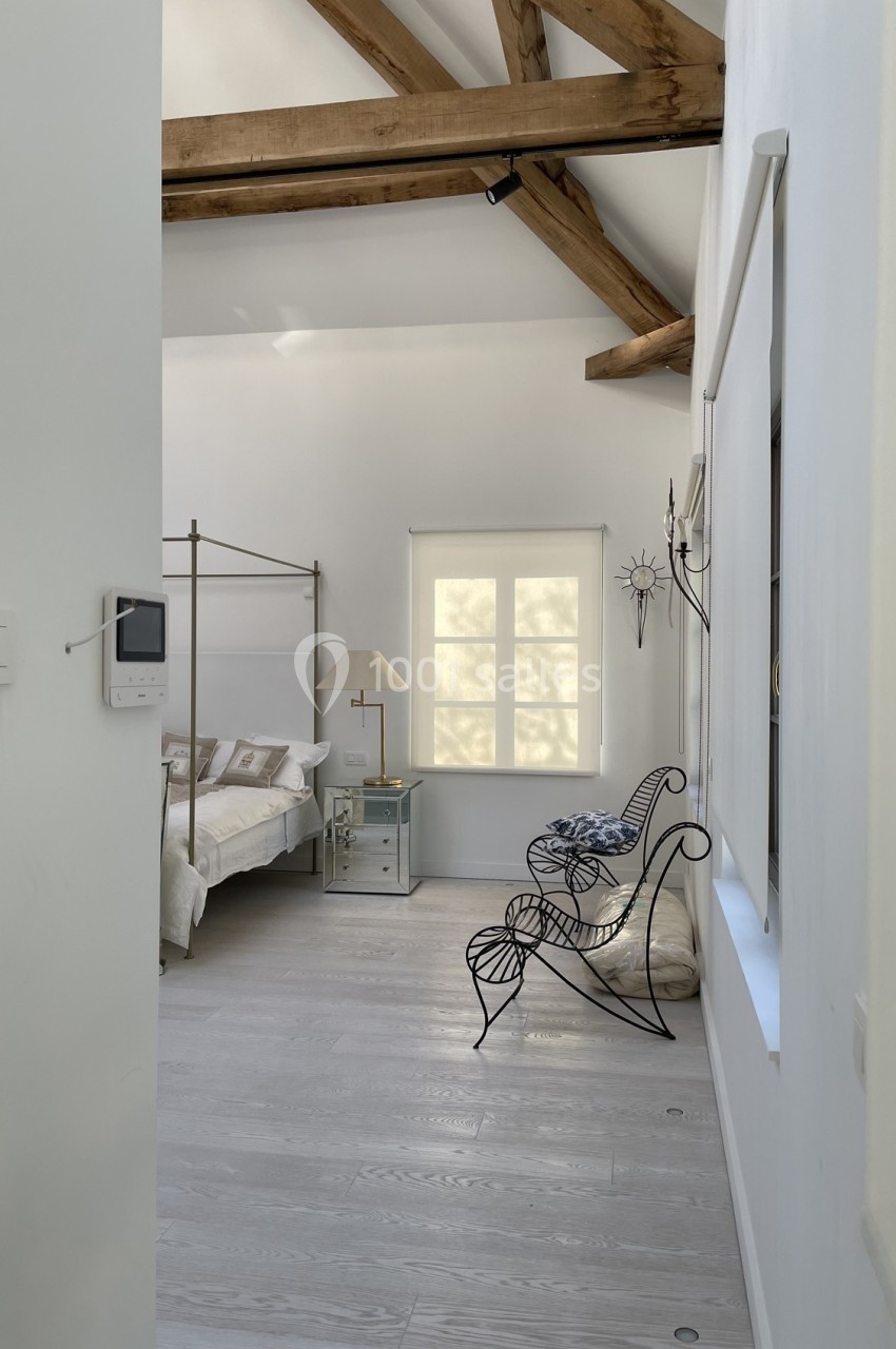 Chambre lumineuse avec lit à baldaquin, fauteuil en métal, poutres apparentes et fenêtre laissant entrer la lumière.