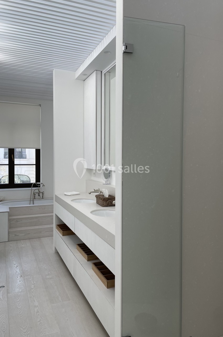 Salle de bain moderne avec double vasque, meubles blancs, sol en bois clair et grande fenêtre avec store.