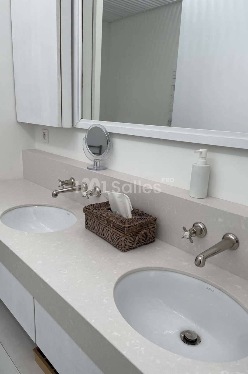 Double vasque blanche avec robinets en métal, miroir mural, panier en osier et distributeur de savon.
