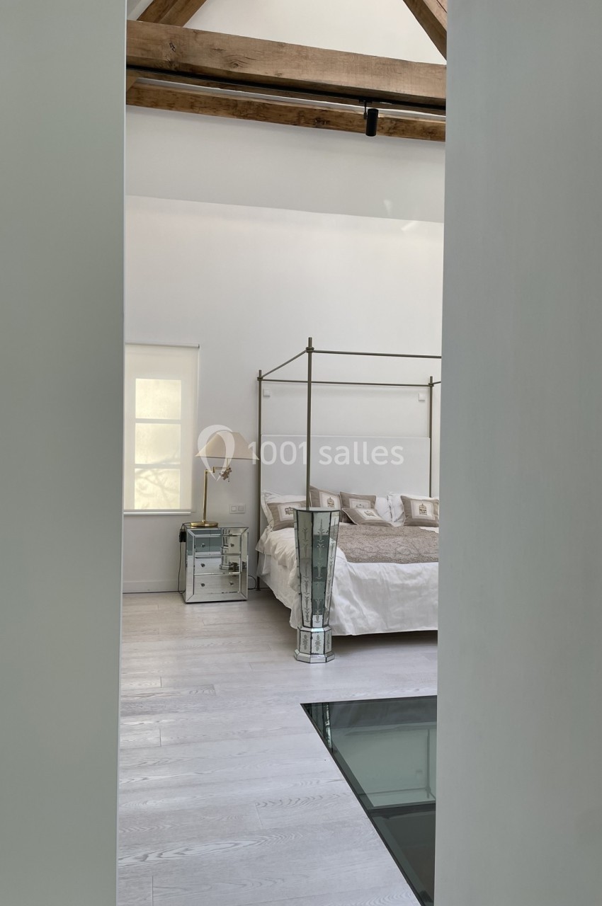 Chambre lumineuse avec lit à baldaquin, parquet clair, poutres apparentes et lampe posée sur une table de chevet.