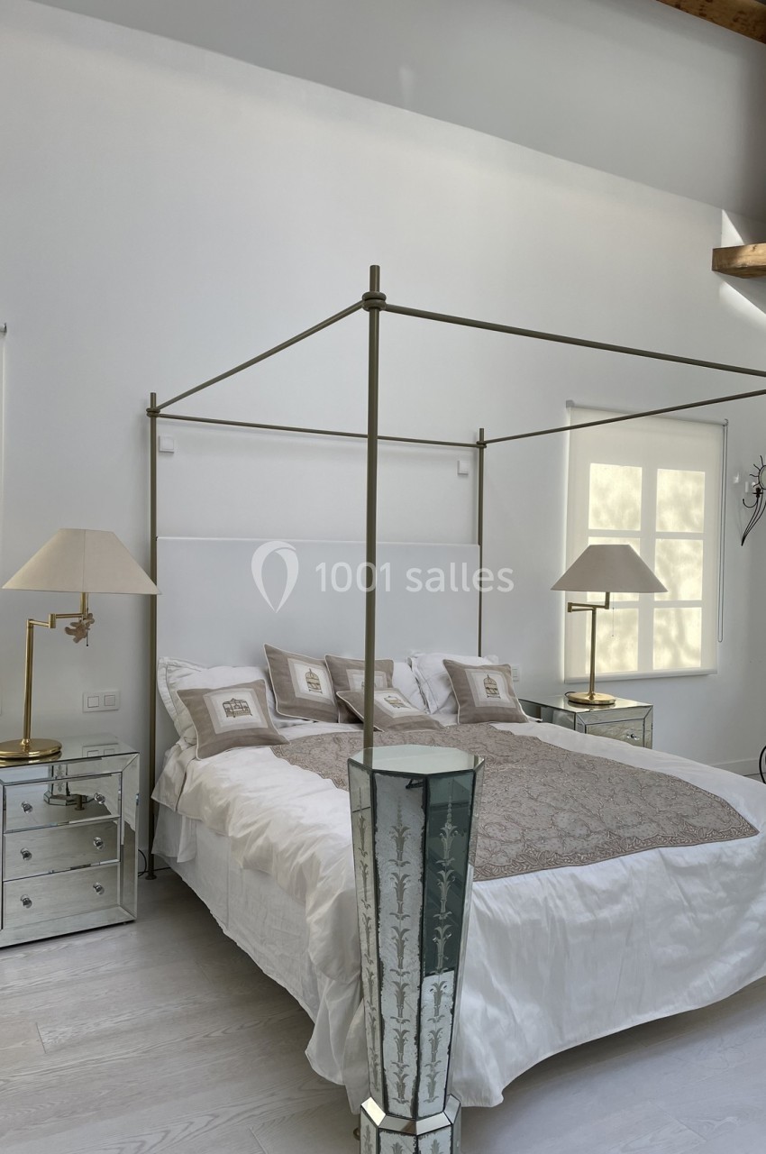 Lit à baldaquin moderne avec draps blancs, tables de chevet en miroir et lampes, dans une chambre lumineuse.